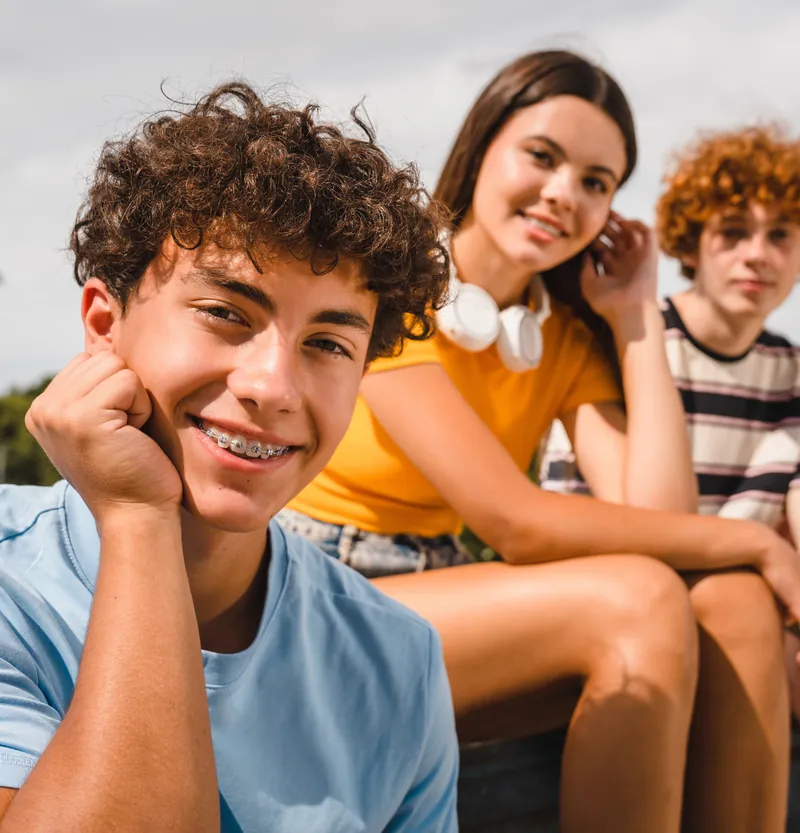 Trois adolescents souriants assis à l’extérieur ; l’un d’eux porte un appareil orthodontique